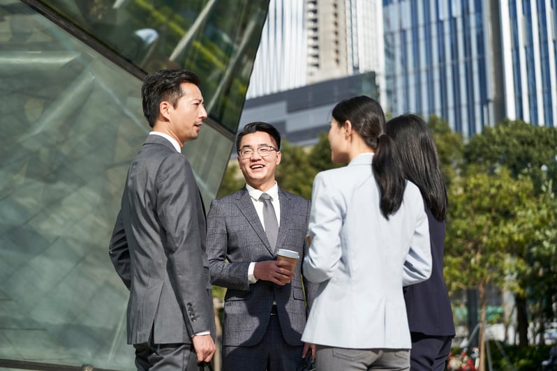 Two male and one female employee(s) facing each other in conversatyion. They are dressed in business attire and standing against a city backdrop with tall commercial buildings. 